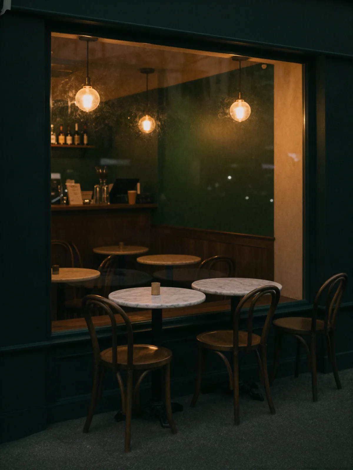 Gece, vitrininden sıcak ışık sızan küçük bir kafe; önünde mermer masalar.